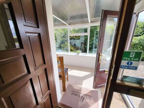 A hotel entrance with a fire exit sign, a wooden door, and a view of the outside greenery. - Farm Cottage at Foot Angus Glens (Kirriemuir)