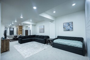 A modern living room with a sectional sofa, a single bed, a small table, and a television. - Spacious and modern basement apartment in serene Mapleton (Mapleton)
