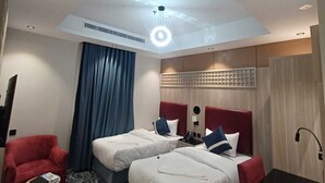 Habitació d'hotel amb dos llits, un butacó vermell, una taula petita, una làmpada al sostre i una catifes amb dibuixos.