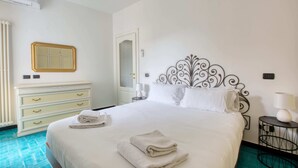 Cama bem arrumada com lençóis brancos, mesa de cabeceira com abajur, cabeceira metálica decorativa e espelho fixado na parede.