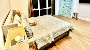 Une chambre à coucher comprenant un lit, une table de chevet, une fenêtre avec des rideaux, un téléviseur fixé au mur et un plancher en bois.