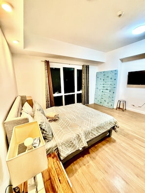 A bedroom with a bed, a nightstand, a window with curtains, a wall-mounted TV, and a wooden floor. - Condo In Abreeza  Family and Spacious (Davao City)