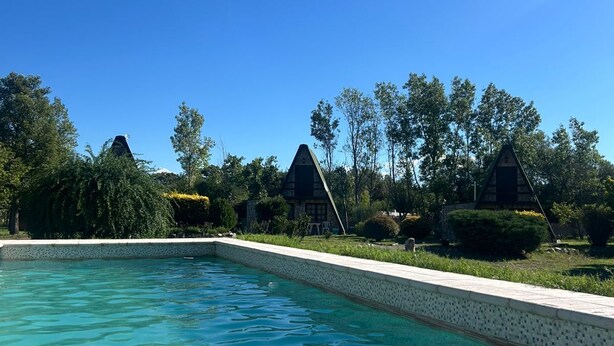 Une piscine aux eaux claires, bordée de carreaux, entourée de verdure et flanquée de deux bâtiments aux toits sombres.