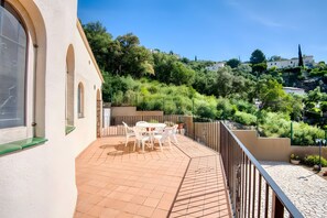 Une terrasse avec une table et des chaises, une balustrade et une vue sur une colline parsemée d’arbres et de bâtiments.
