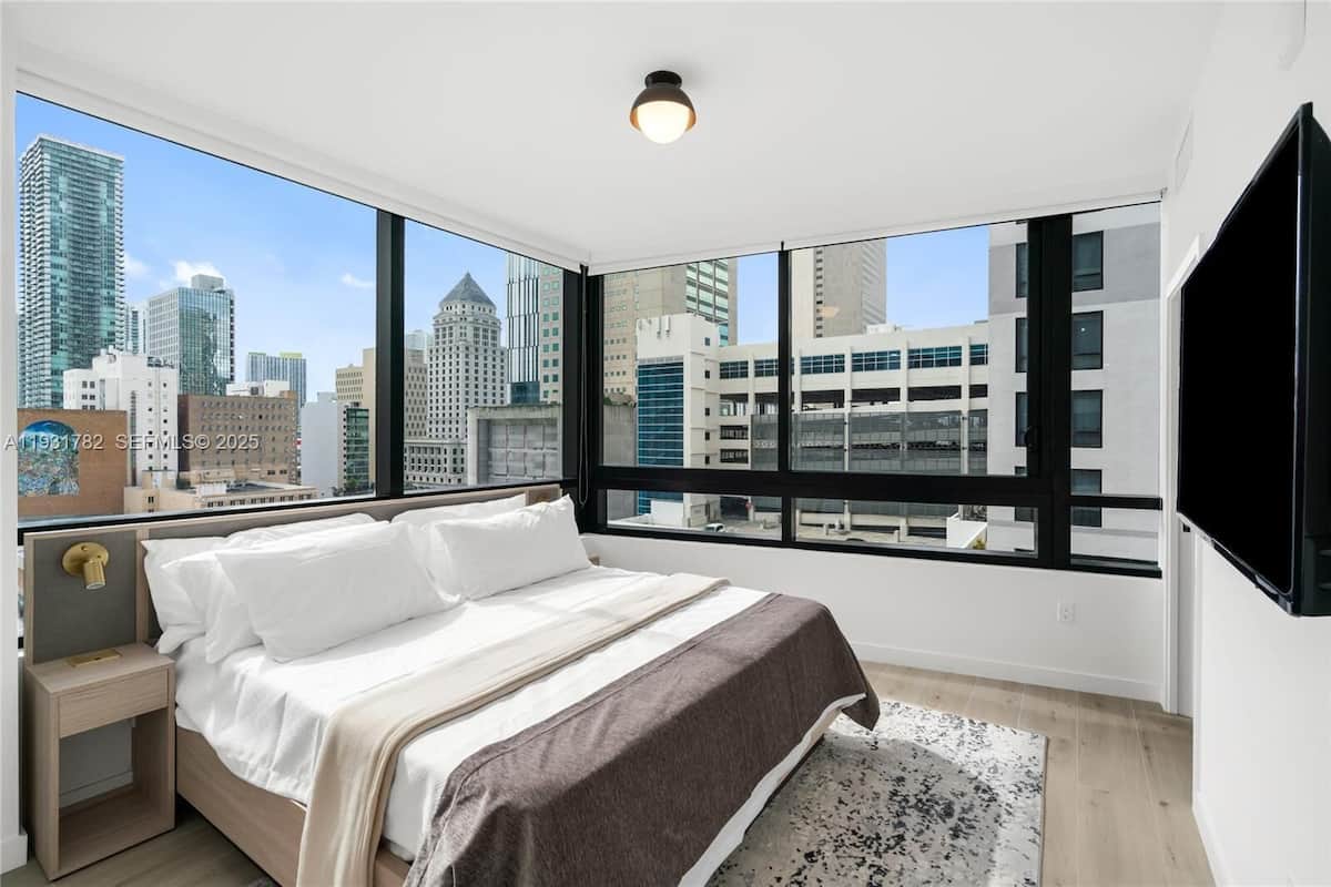 A bedroom with a bed, a nightstand, a large window with a city view, and a ceiling light.