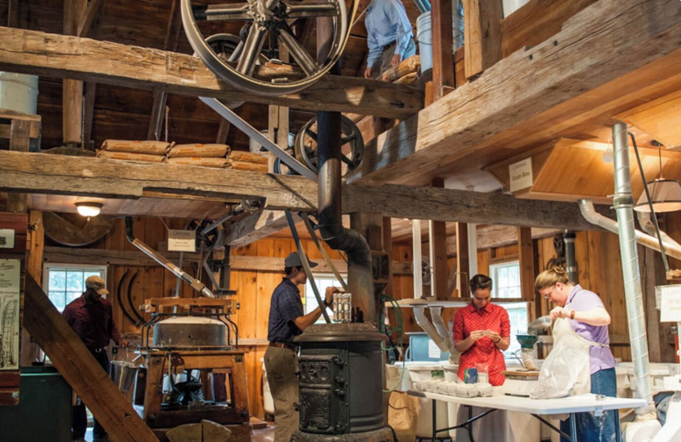 Un intérieur rustique avec des poutres apparentes, un grand poêle noir et une table autour de laquelle des personnes travaillent.