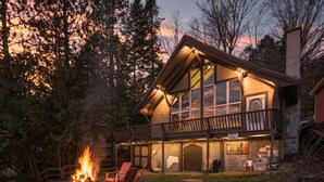 Un chalet à deux étages doté d’un foyer extérieur et de sièges en plein air, le tout entouré d’arbres.