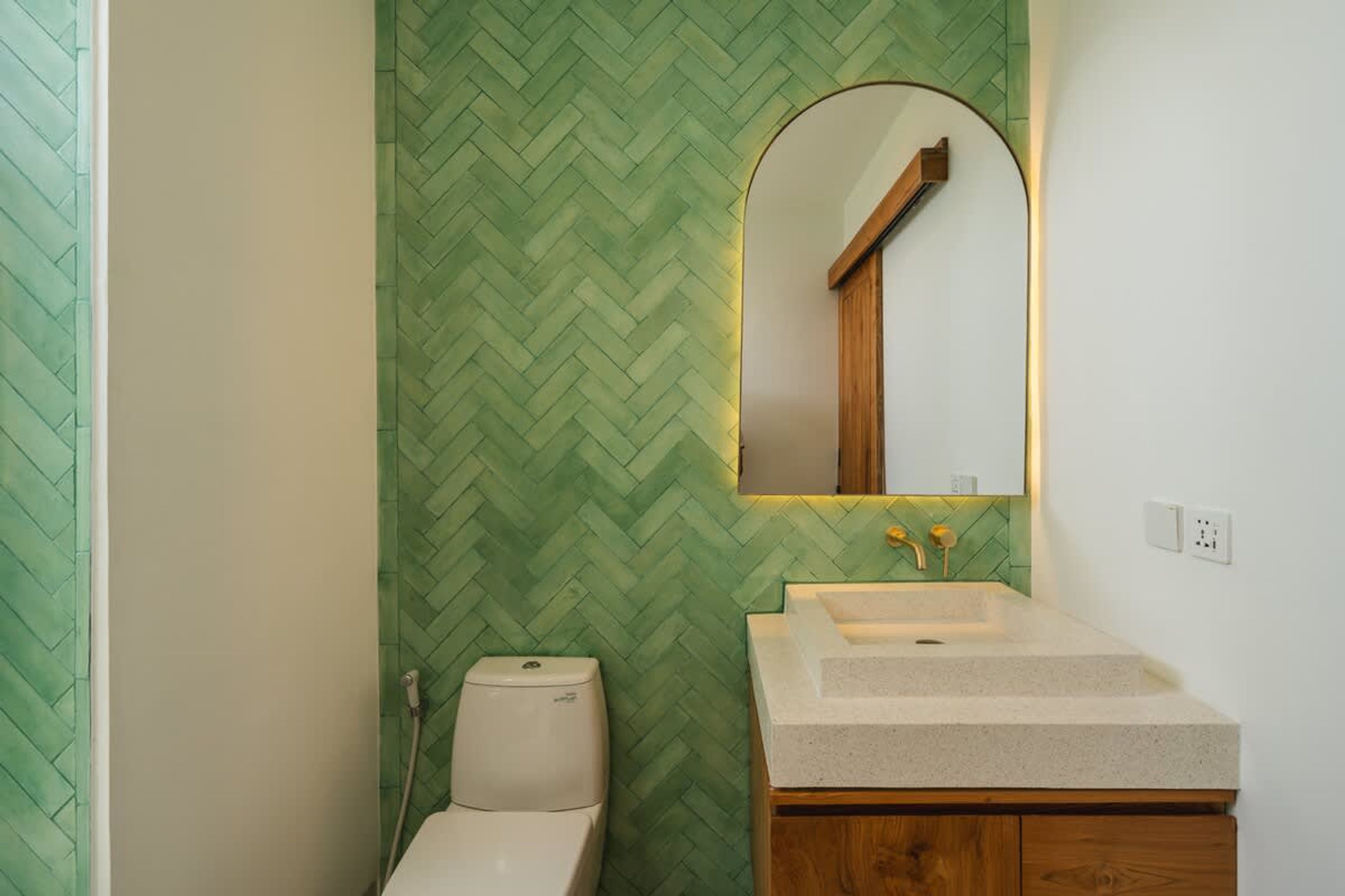 A bathroom with a green herringbone-patterned wall, a white toilet, a wooden vanity with a sink, and a mirror with a wooden frame.