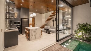A modern kitchen with a marble island, a dining area with chairs, a staircase, and a glass-enclosed living space with a pool. - Rocca Bella Loft 3 in Canggu (Canggu)