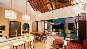 Eine moderne Küche mit einer zentralen Kochinsel, Barhocker, einem Essbereich mit Tisch und Stühlen sowie einer Sitzgelegenheit am Pool mit einer roten Couch und runden Tischen.