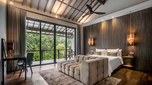 Une chambre moderne dotée d’un grand lit, d’un coin salon, d’un bureau et offrant une vue sur la verdure grâce à de grandes fenêtres.