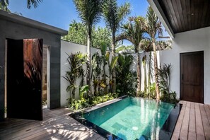 Ein moderner Poolbereich mit Holzterrasse, umgeben von tropischen Pflanzen und einer weißen Wand.