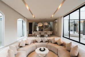 A modern living room with a curved sofa, a dining area with a round table, and large windows. - Villa Sayuri in Kecamatan Kuta Utara (Kecamatan Kuta Utara)