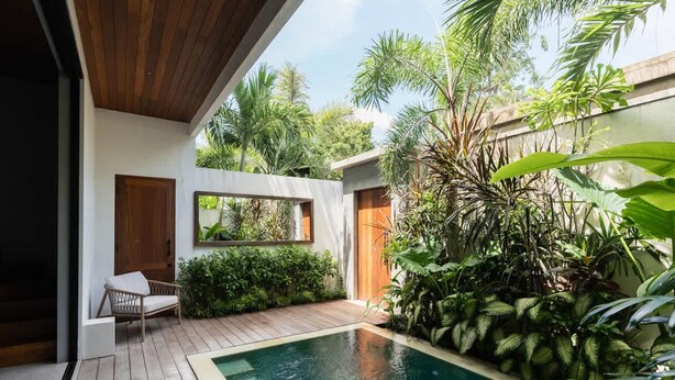 Uma área moderna de piscina externa com deck de madeira, uma espreguiçadeira e exuberante vegetação tropical.