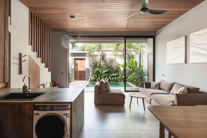 A modern kitchen with a washing machine, a dining area with a table and chairs, a sofa, and a view of a swimming pool and garden. - Umayam Sanctuary in Pererenan (Pererenan)