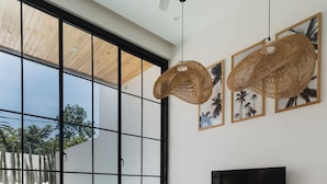 Ein modernes Wohnzimmer mit großem Fenster, weißen Sofas, einem runden Korbtisch und einem an der Wand befestigten Flachbildfernseher.