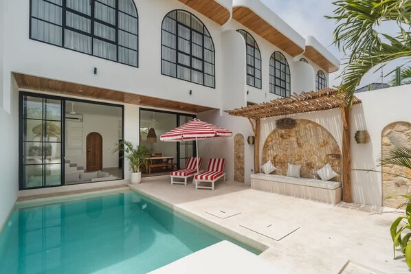 Une maison moderne dotée d’une piscine, de grandes fenêtres et d’un patio aménagé avec des chaises longues et un parasol.