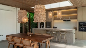 Een moderne keuken met een centraal kookeiland, een houten eettafel en rieten hanglampen.