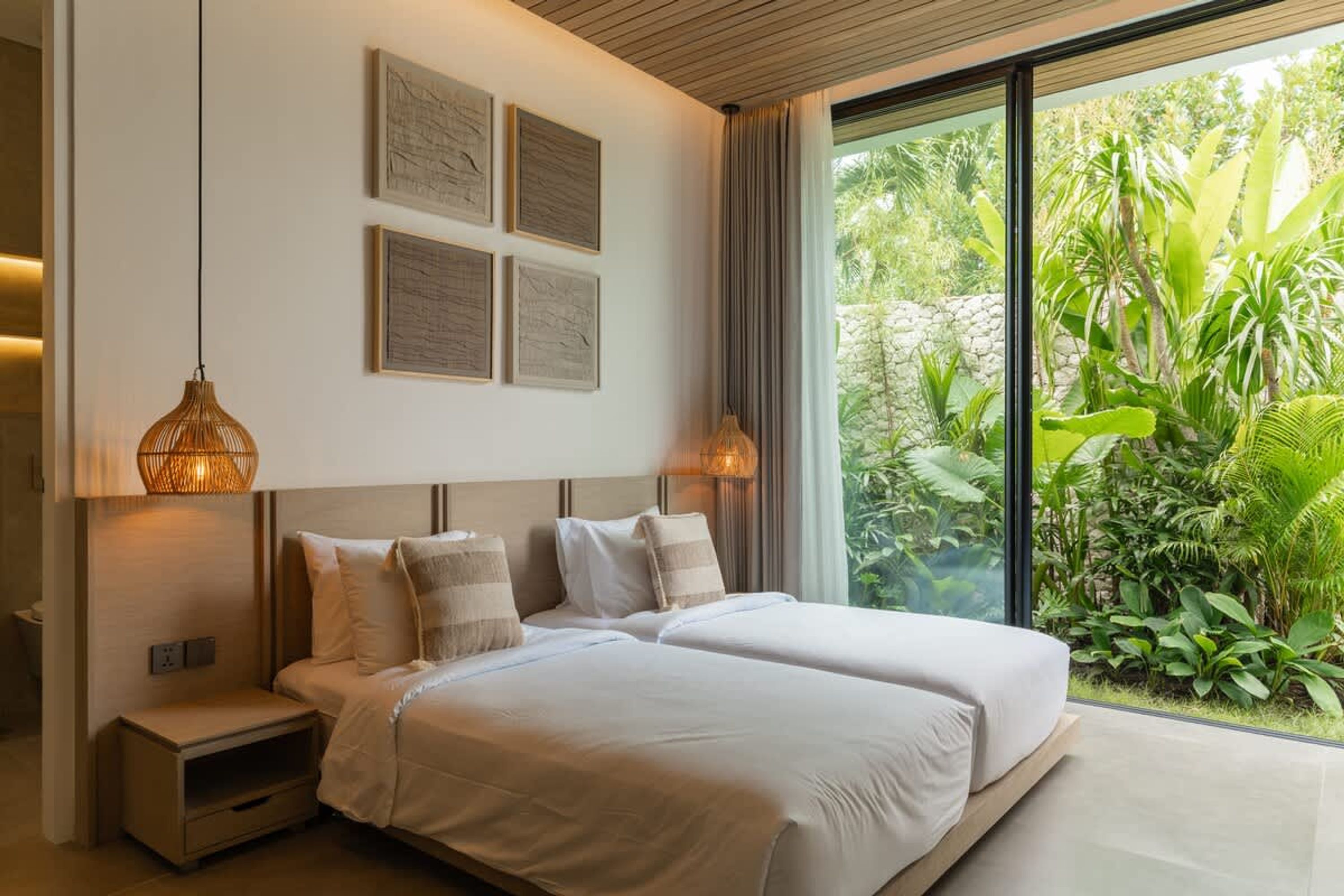 A bedroom with two beds, a nightstand, a wall-mounted lamp, and a large window with a view of greenery.