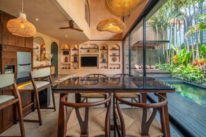 Un comedor moderno con una mesa de vidrio, sillas de madera y vistas a una piscina y plantas tropicales.