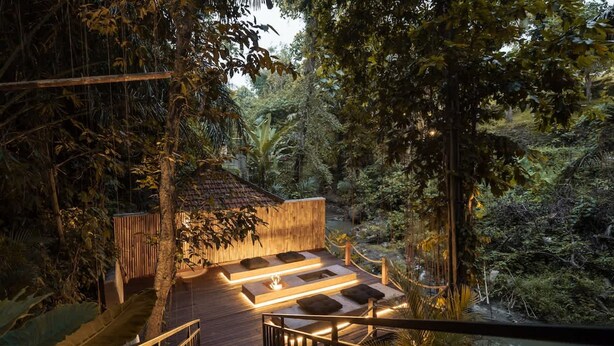 Una terraza de madera con un hoyo de fuego, rodeada de follaje denso y una estructura de madera.