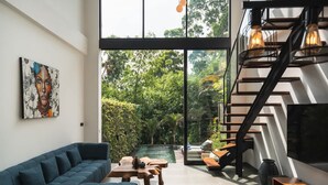 Un interior moderno con sofá, mesa de madera, escalera y vistas a un jardín exuberante.