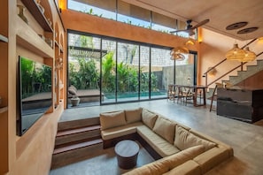 A modern living room with a large sectional sofa, a dining area, and a staircase leading to an outdoor space. - The Mystik Loft Serenity (Pererenan)