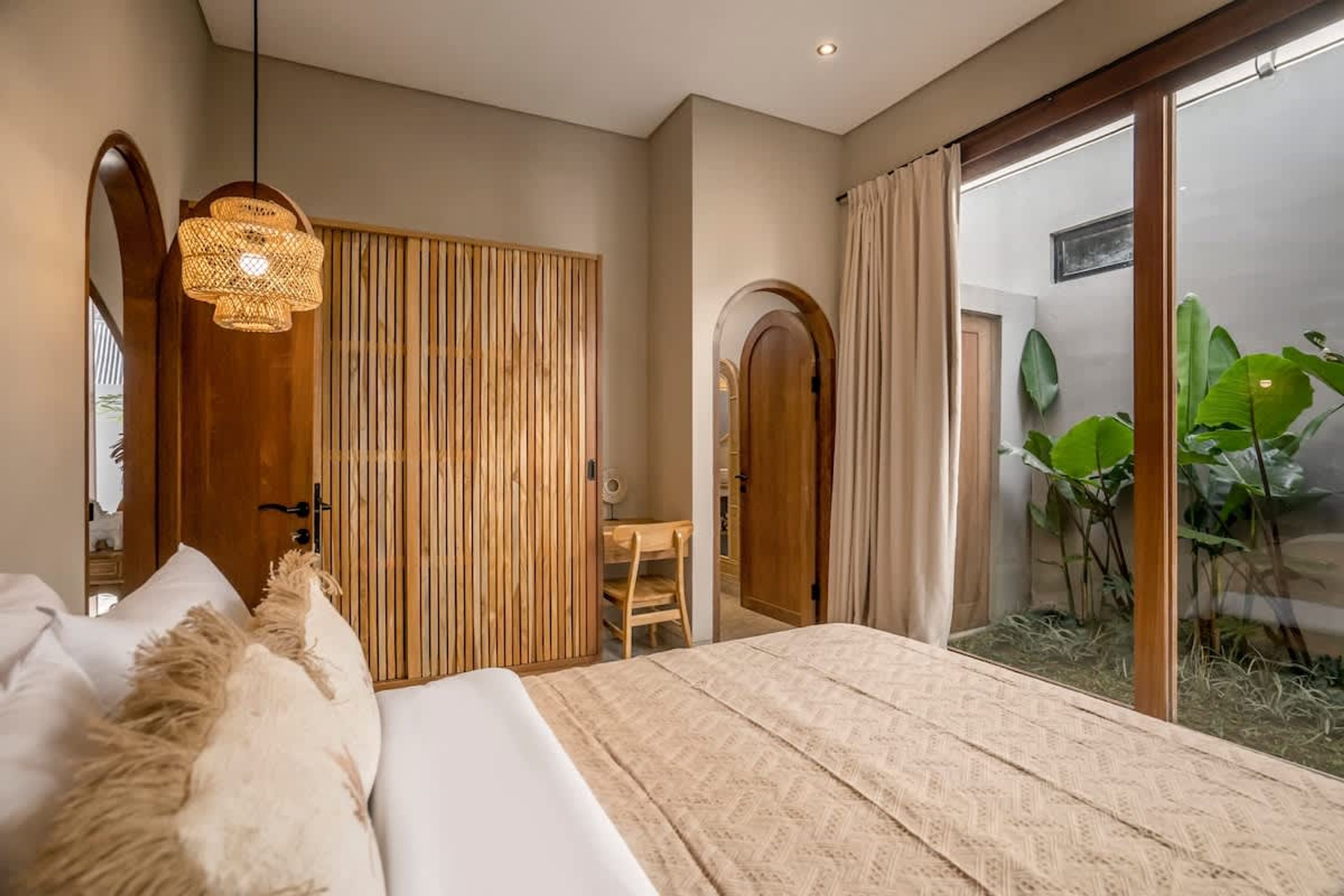 A bedroom with a bed, a wooden wardrobe, a desk, and a sliding glass door leading to a balcony with plants.