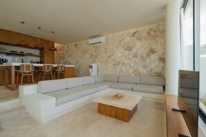 A modern living room with a sectional sofa, a wooden coffee table, and a kitchen area with wooden cabinets and pendant lights. - Villa Beyond  Seashell (South Kuta)