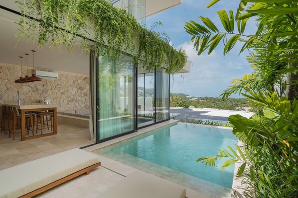 A modern pool area with a glass-enclosed infinity pool, a wooden dining set, and lush greenery. - Villa Beyond  Seashell (South Kuta)