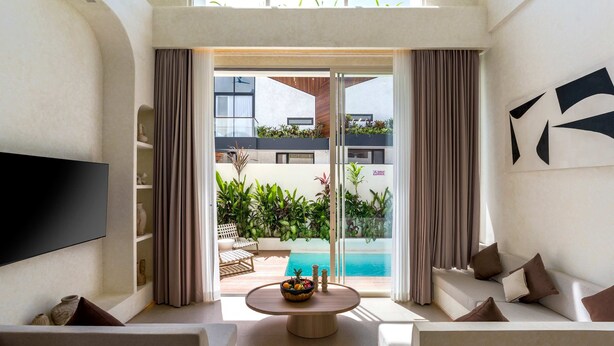 Ein modernes Wohnzimmer mit einem Flachbildfernseher, einem runden Couchtisch mit Obstschale, einer Couch und Blick auf einen Swimmingpool und Grünflächen im Freien.