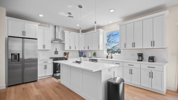 A modern kitchen with white cabinetry, a stainless steel refrigerator, a central island, pendant lighting, and a window providing natural light.