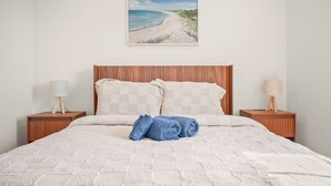 A neatly made bed with a checkered pillow, two bedside tables with lamps, and a beach painting on the wall.