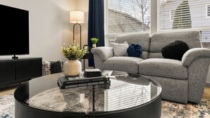 A modern living room with a grey sofa, a round coffee table with books, a TV, and a lamp. - Quiet Boise Retreat|Hot Tub•Game Room• 4BR (Boise)