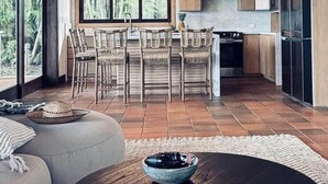Een moderne keuken met houten kastjes, een eethoek met rieten stoelen en een groot raam met uitzicht op groen.