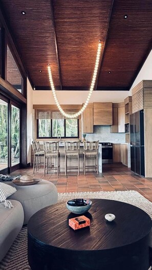 A modern kitchen with wooden cabinets, a dining area with wicker chairs, and a large window overlooking greenery. - Villa Las Brisas: Ocean View and Walk to the Beach (Sámara)