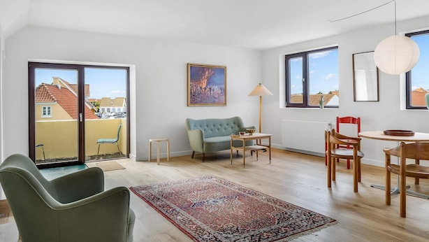 A modern living room with a sofa, a dining table, and chairs. There are large windows and a view of buildings outside. - 4 Star Holiday Home in Lokken (Lokken)
