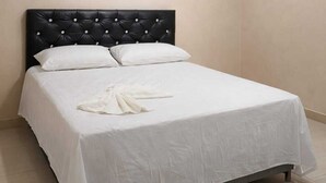 Una cama individual con cabecera negra y ropa de cama blanca en una habitación con paredes lisas y suelo de baldosas.