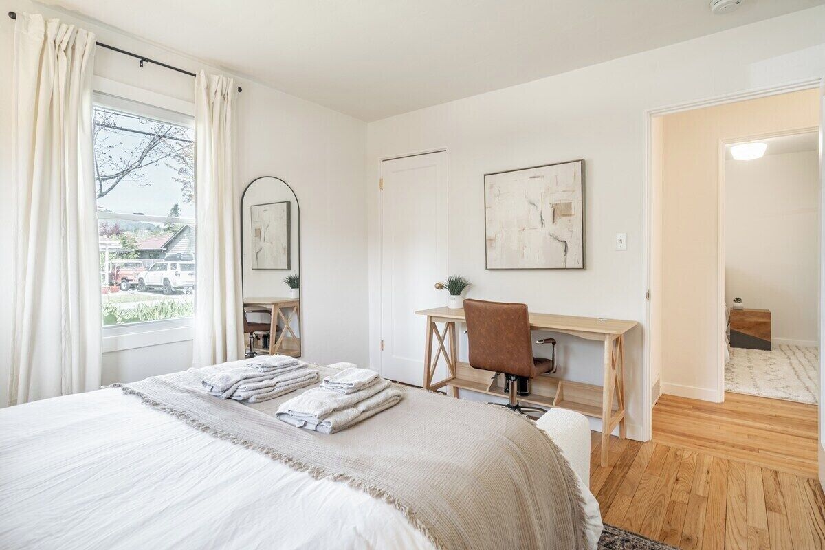 A bedroom with a bed, a desk, and a chair. There is a window with curtains, a door, and a view of the outside.