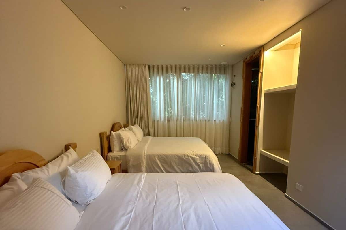 Una habitación de hotel con dos camas, un ventanal grande con cortinas, un armario empotrado y un espejo.