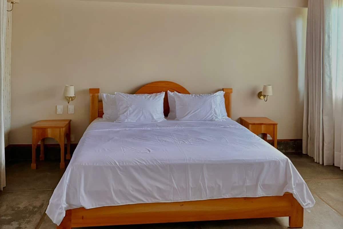 Una cama con ropa de cama blanca, cabecero de madera, dos mesitas de noche con lámparas y techo de madera.