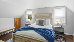 Une chambre avec un plafond en pente, un lit recouvert d’une couverture bleue, deux tables de chevet avec des lampes, un banc en bois et une armoire.