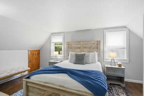 A bedroom with a sloped ceiling, a wooden headboard, a bed with a blue blanket, two bedside tables with lamps, a bench, and a wardrobe.