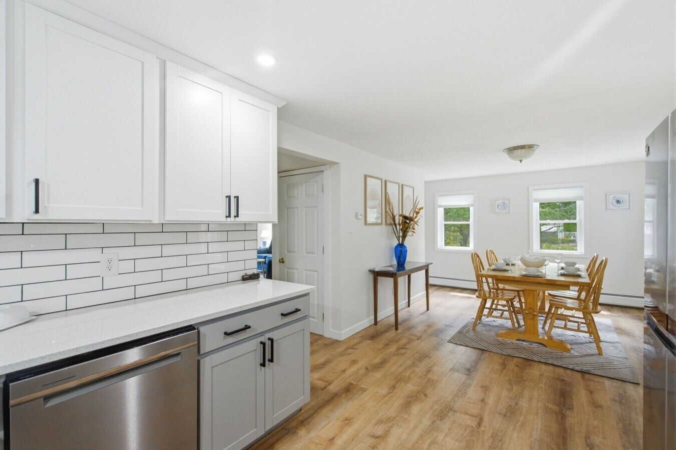 Una cocina con armarios blancos, un lavavajillas de acero inoxidable y un comedor con mesa y sillas de madera.