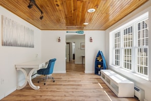 A room with a wooden ceiling, a desk with a chair, a window with blinds, and a blue shelf with a vase.