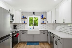 Una cocina moderna con armarios blancos, un refrigerador de acero inoxidable, horno empotrado y ventana con vista a follaje.