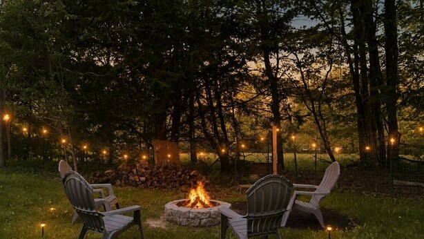 Un espace extérieur aménagé avec des chaises et un foyer, entouré d’arbres et illuminé de guirlandes lumineuses.
