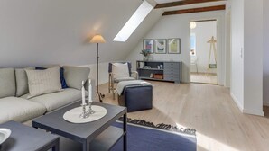 Un loft moderno con vigas de madera a la vista, un sofá gris, una pequeña mesa con velas, una lámpara de pie y vistas de la ciudad a través de un tragaluz.