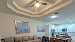 Una sala de estar moderna con un sofá modular, una mesa de comedor con sillas y una zona de cocina con armarios y electrodomésticos blancos.