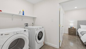 Ein Waschraum mit zwei Whirlpool-Waschmaschinen, einer Ablage mit Reinigungsmitteln und ein Schlafzimmer mit Bett und Nachttisch.